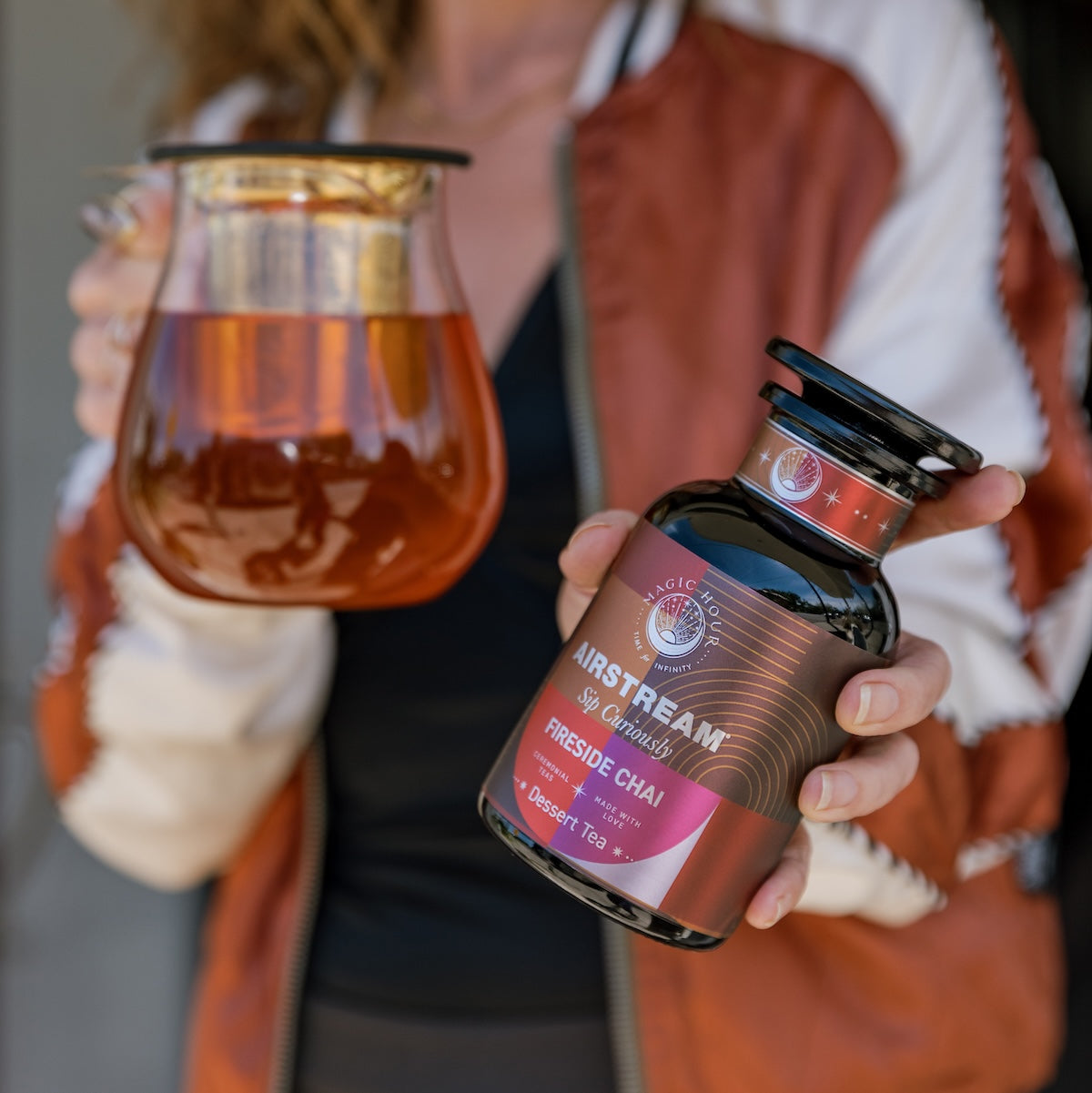 A person wearing a red and white jacket holds a glass pitcher of tea in one hand and a container labeled &quot;Magic Hour - Fireside Chai: Warming Rooibos Blend for Creating Connection&quot; in the other. The label, which features a red and black design, highlights that it&#39;s a caffeine-free spiced herbal chai packed with antioxidants including vitamin C.