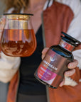 A person wearing a red and white jacket holds a glass pitcher of tea in one hand and a container labeled "Magic Hour - Fireside Chai: Warming Rooibos Blend for Creating Connection" in the other. The label, which features a red and black design, highlights that it's a caffeine-free spiced herbal chai packed with antioxidants including vitamin C.