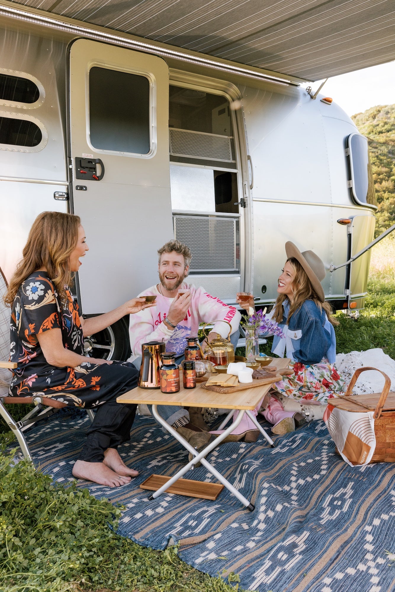 Three people sit on a patterned outdoor rug under the awning of an Airstream Camper, enjoying a picnic. They are surrounded by various foods, drinks, and loose leaf tea on a low table. The setting is sunny with a grassy landscape in the background. All three are smiling and interacting.