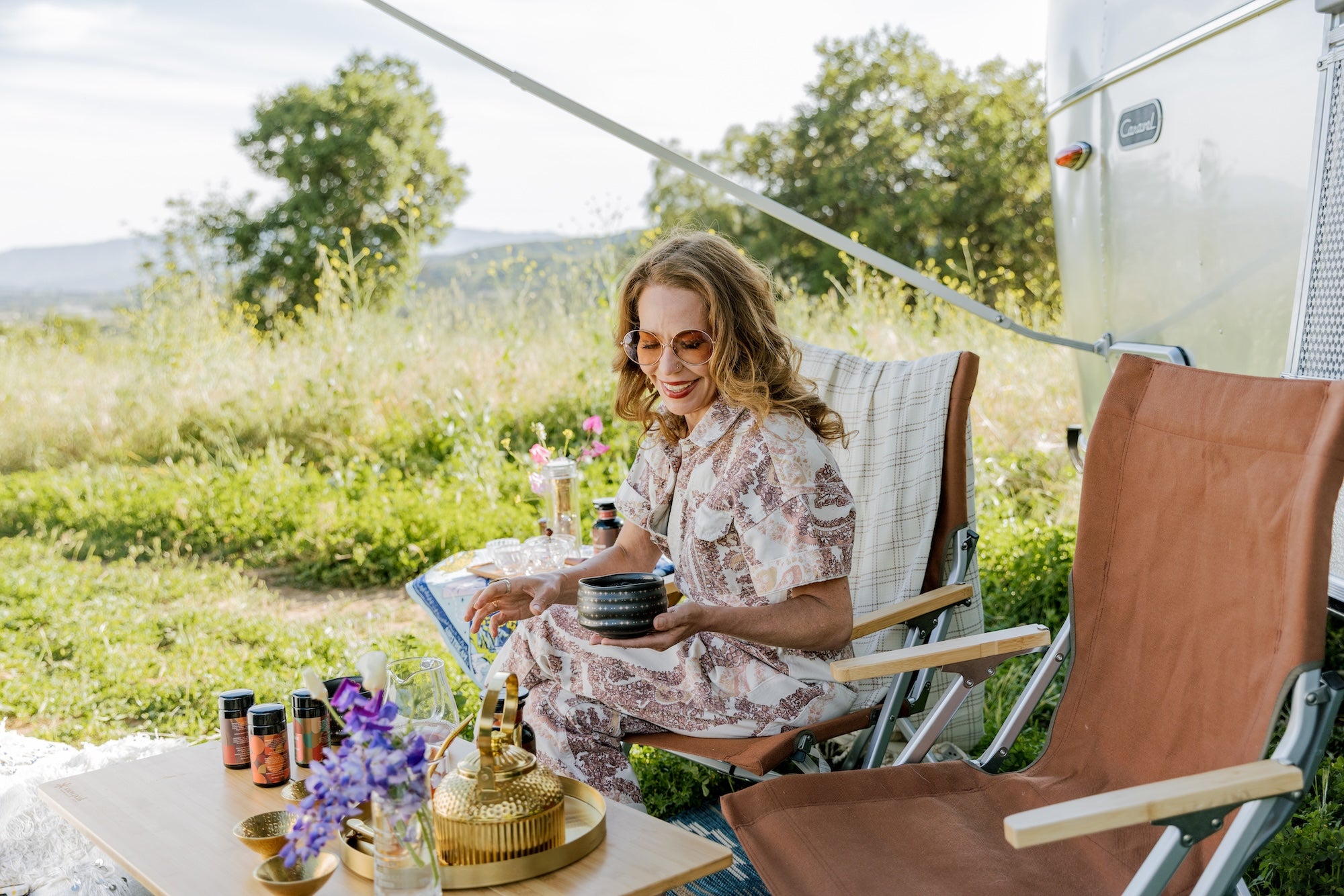 A woman with long hair and sunglasses, wearing a patterned dress, is sitting on a chair next to a table with various items outdoors. She is holding a small bowl. Behind her, there is an Airstream trailer and lush greenery under a clear sky.