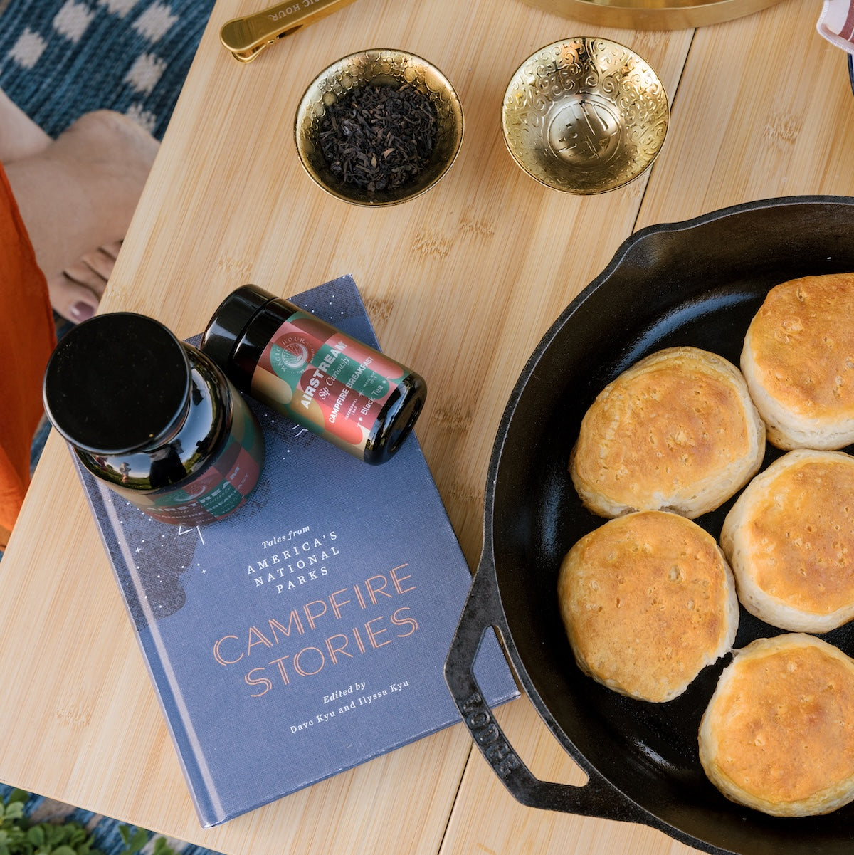 Campfire Breakfast Black Loose Leaf Tea on a wooden table with a campfire stories book and a cast iron skillet with biscuits. 