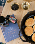 Campfire Breakfast Black Loose Leaf Tea on a wooden table with a campfire stories book and a cast iron skillet with biscuits. 