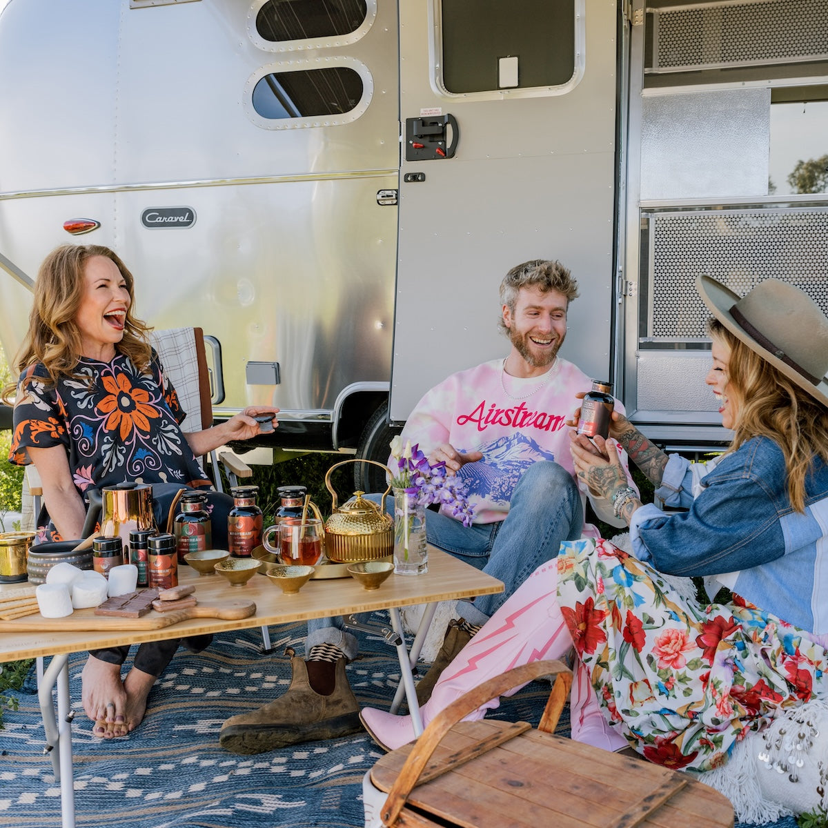 Three people sit and laugh by an Airstream trailer. They are surrounded by skincare products and a kettle on a table. The background includes part of the trailer and open trailer doors.