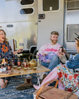 Three people sit and laugh by an Airstream trailer. They are surrounded by skincare products and a kettle on a table. The background includes part of the trailer and open trailer doors.