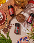 Top view of a cozy tea setup featuring the Sip Curiously Gift Set from Magic Hour, including a wooden board, a gold teapot, three tins of loose tea, an exclusive blends latte in a red cup, the Midas Touch Tea strainer, assorted flowers, playing cards, and a red-and-white patterned cloth. Scattered tea leaves add texture to the scene.