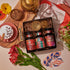 A cozy scene features the Magic Hour's Sip Curiously Gift Set with three tins labeled S'mores, Ginger Breadhouse, and Fireside Chai. Surrounding the box are a bowl of frothy milk with chocolate chips, flowers, a teapot, cards, chocolate shavings, and a colorful cloth.