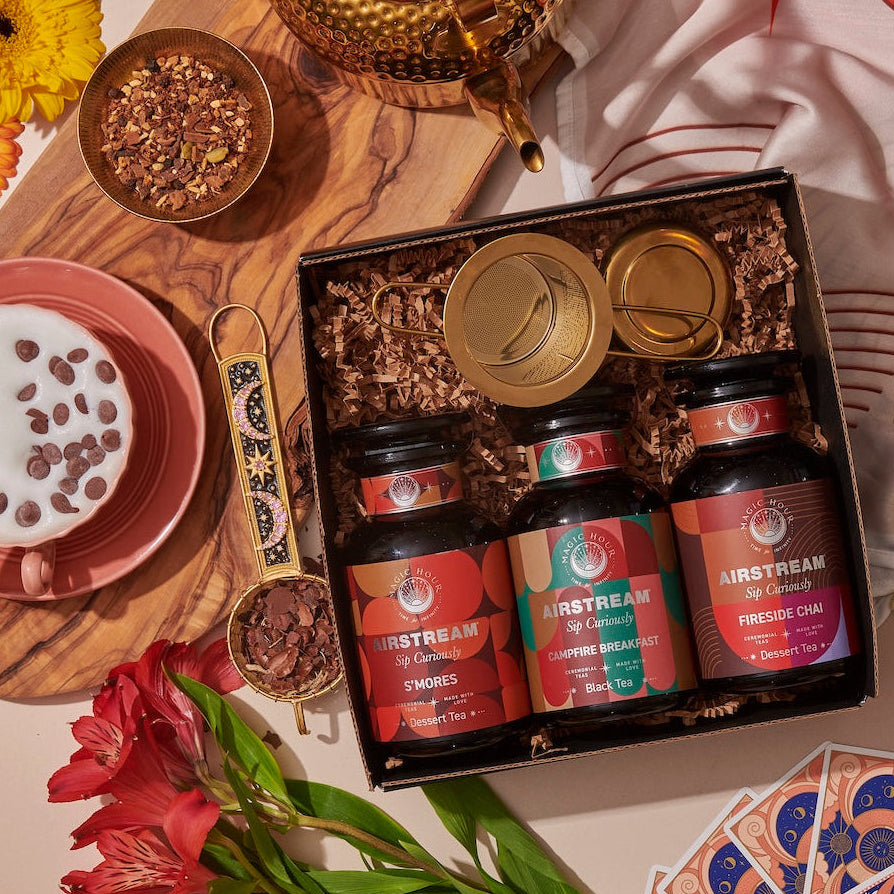 A cozy scene features the Magic Hour's Sip Curiously Gift Set with three tins labeled S'mores, Ginger Breadhouse, and Fireside Chai. Surrounding the box are a bowl of frothy milk with chocolate chips, flowers, a teapot, cards, chocolate shavings, and a colorful cloth.