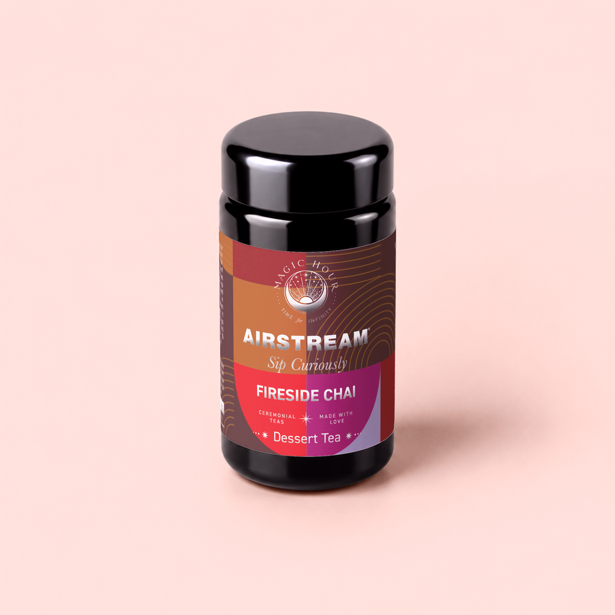 A jar of Fireside Chai: Warming Rooibos Blend for Creating Connection, a caffeine-free spiced herbal dessert tea by Magic Hour, features a black lid and warm-toned labels with the words “Sip Curiously” on a soft pink background. This antioxidant-rich rooibos blend invites cozy moments with every cup.