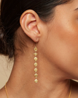 A woman with her hair in a bun is wearing the Aligned in Harmony Earrings, which are long, 18kt gold-plated drop earrings adorned with multiple small, intricate charms. She has warm-toned, glowing skin and the background is a soft beige color, creating a harmonious balance in the image.