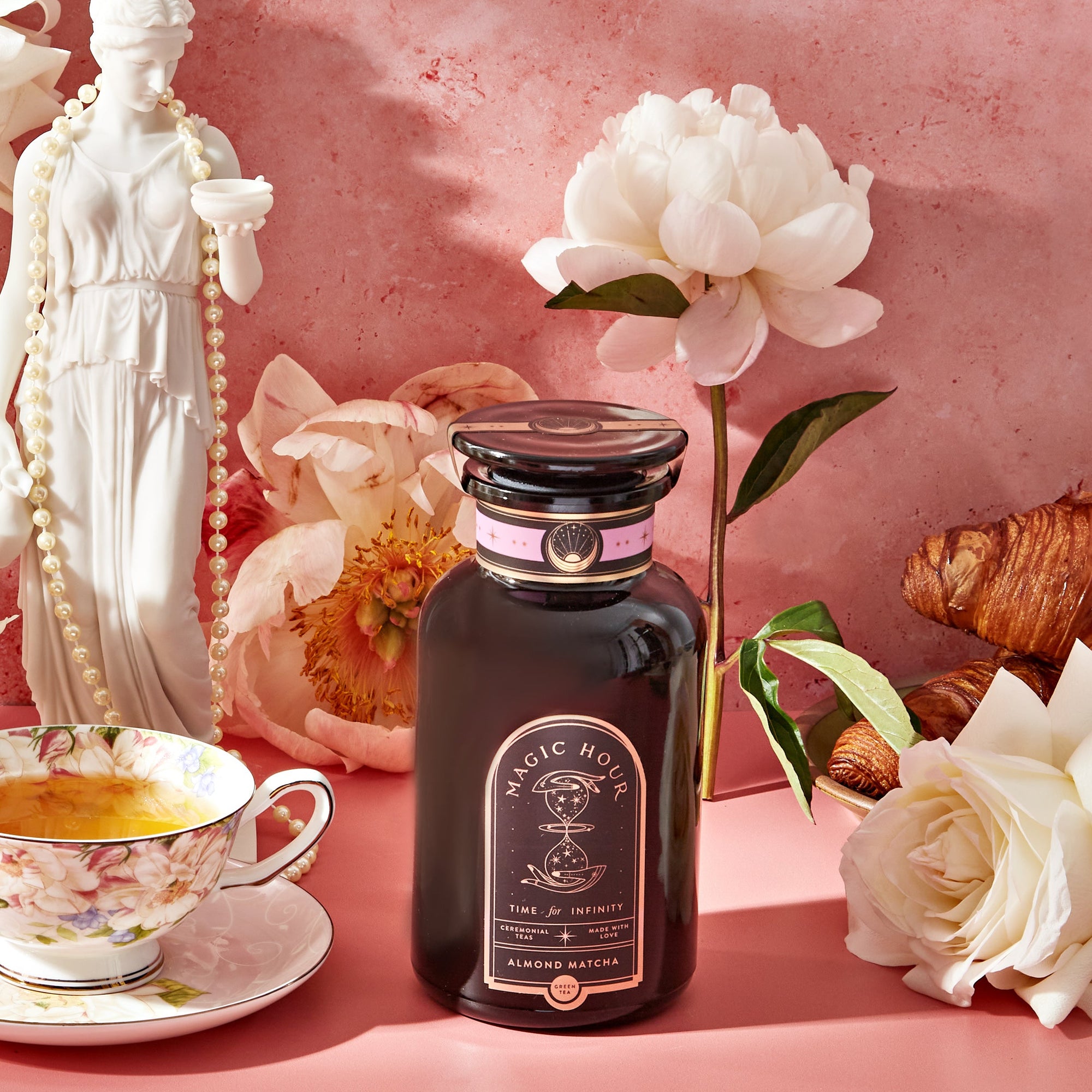 A jar of honey rests on a pink surface with white flowers, a floral teacup and saucer filled with Almond Matcha Green Tea for Joy, a croissant, and a white statue of a woman set against a pink textured background.