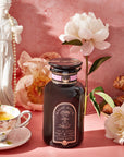 A jar of honey rests on a pink surface with white flowers, a floral teacup and saucer filled with Almond Matcha Green Tea for Joy, a croissant, and a white statue of a woman set against a pink textured background.