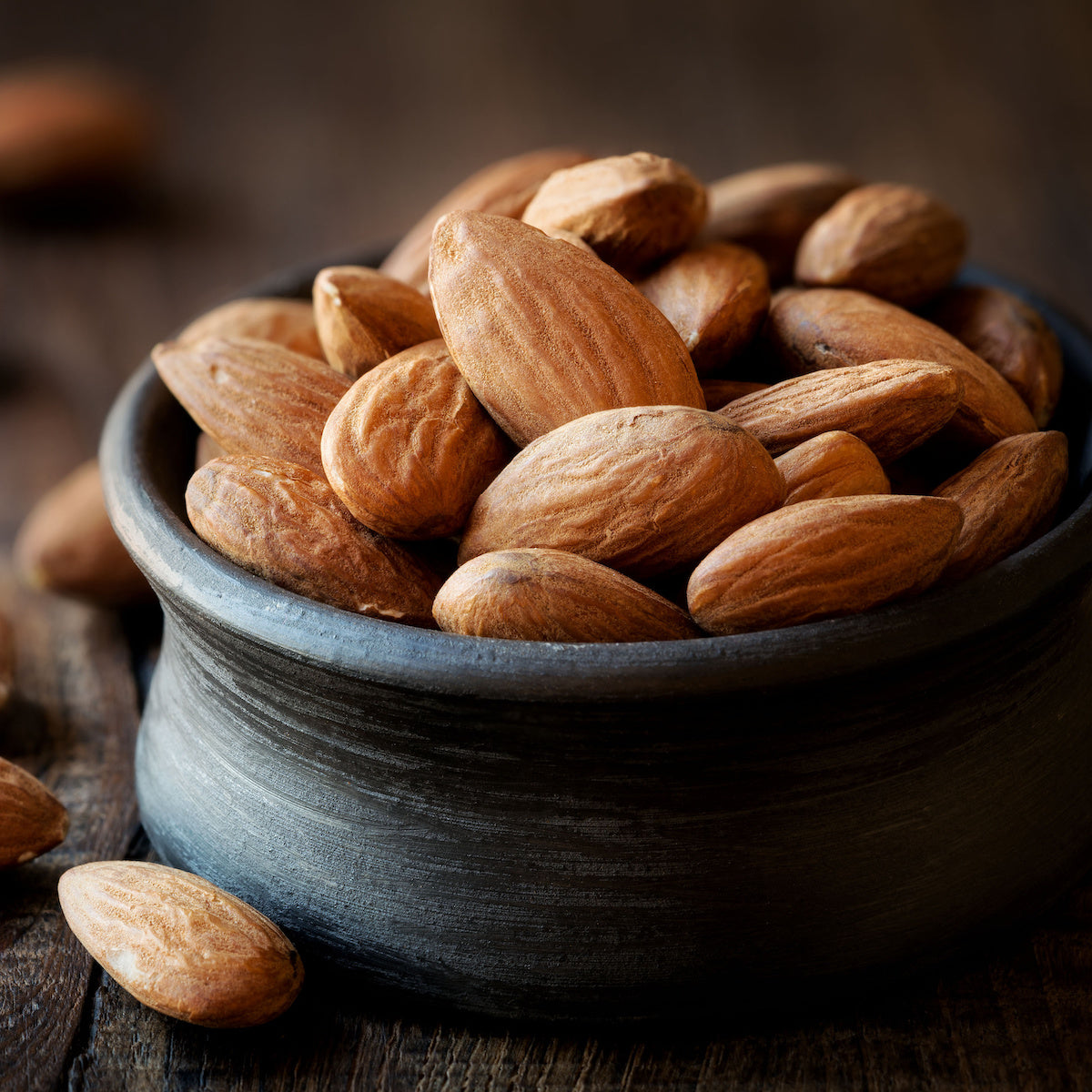 A ceramic bowl filled with unshelled almonds is placed on a wooden surface. The almonds are smooth and brown, resting in a rustic, black bowl. A few almonds are scattered around the bowl, adding to the natural, earthy ambiance of the scene.