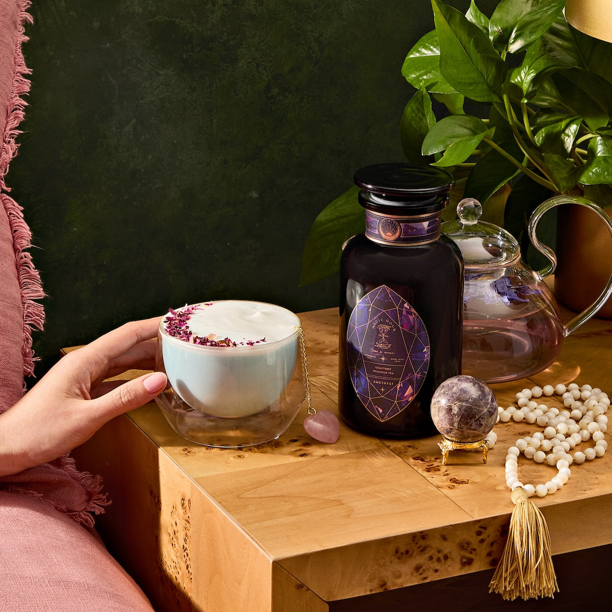 A hand holds a mug of pink petal-topped Amethyst: Strawberry-Passionfruit Gemstone Wellness Tea beside a wooden table. On the table is a geometrically designed dark bottle, amethyst beads for clarity, a crystal sphere, and a teapot with lush green leaves enhancing intuition.