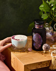 A hand holds a mug of pink petal-topped Amethyst: Strawberry-Passionfruit Gemstone Wellness Tea beside a wooden table. On the table is a geometrically designed dark bottle, amethyst beads for clarity, a crystal sphere, and a teapot with lush green leaves enhancing intuition.