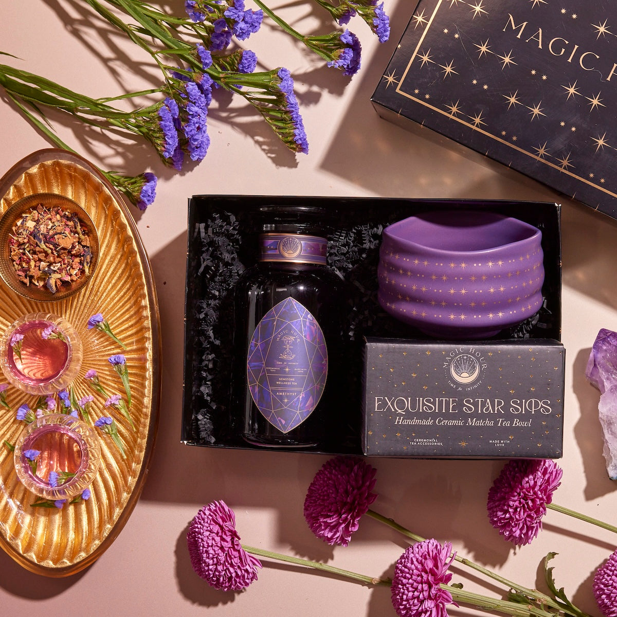 A flat lay image showcases an "Amethyst Gemstone Tea Gift Set," featuring a bottle of Amethyst Tea, a purple ceramic mug, and elements of spiritual insight. The arrangement is adorned with flowers, a crystal promoting Third Eye Chakra harmony, and a golden tray displaying tea leaves along with small bowls.