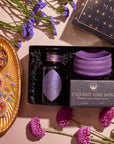 A flat lay image showcases an "Amethyst Gemstone Tea Gift Set," featuring a bottle of Amethyst Tea, a purple ceramic mug, and elements of spiritual insight. The arrangement is adorned with flowers, a crystal promoting Third Eye Chakra harmony, and a golden tray displaying tea leaves along with small bowls.