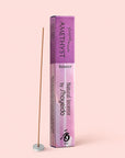 A box of Natural Incense : Amethyst, offering silky notes of sandalwood and cinnamon, stands upright next to a single incense stick in a small white holder, set against a light pink background.