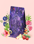 A vibrant purple Amethyst: Strawberry-Passionfruit Wellness Tea Refill Pouch, adorned with a geometric crystal design that reflects the spirit of Amethyst tea and holistic wellness, sits amid illustrated flowers, strawberries, apple slices, and green leaves—all displayed on a soft pink background.