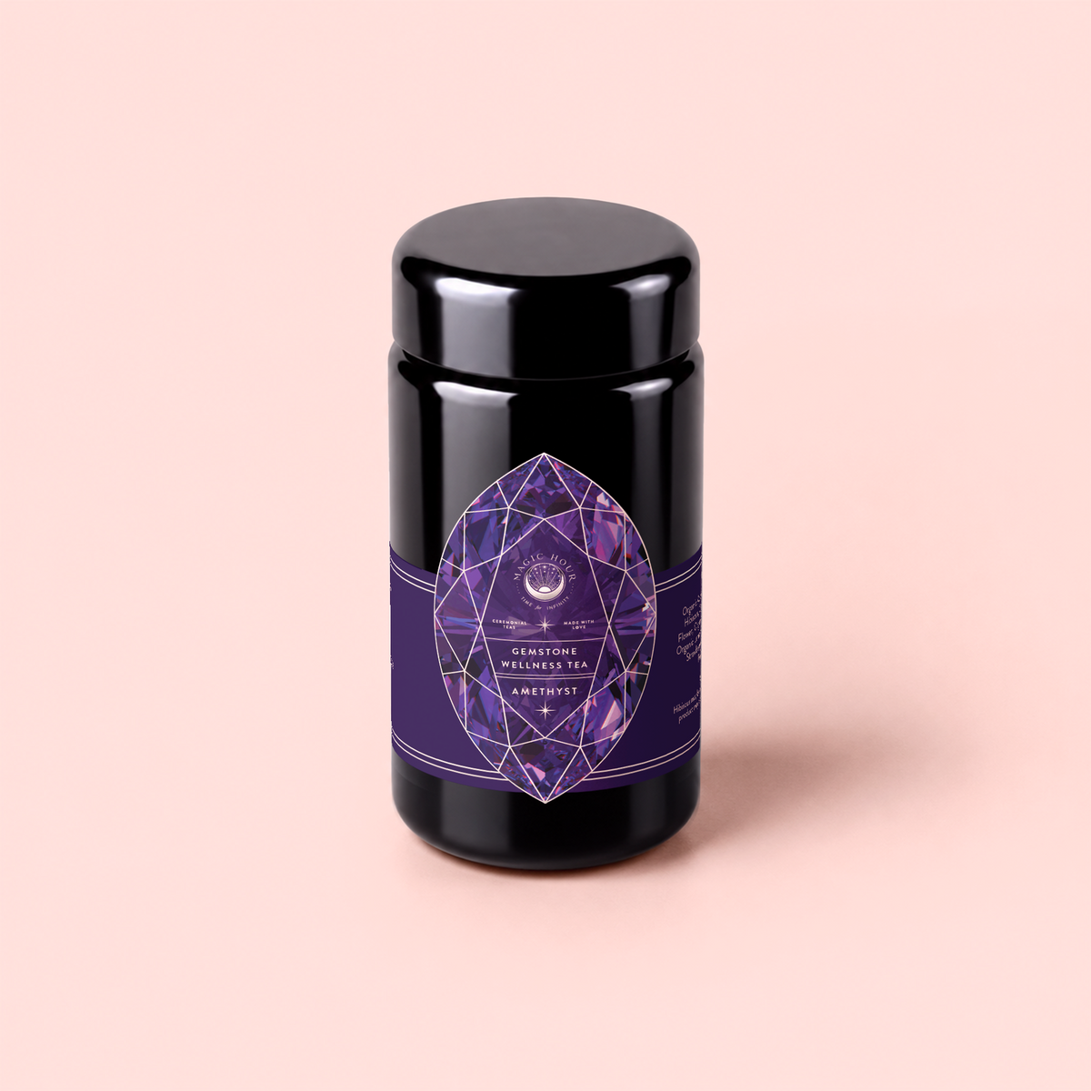 A black jar adorned with a purple geometric crystal label reading “Amethyst: Strawberry-Passionfruit Gemstone Wellness Tea” rests on a pale pink background, evoking spiritual wellness and the calming aura of gemstone-infused tea.