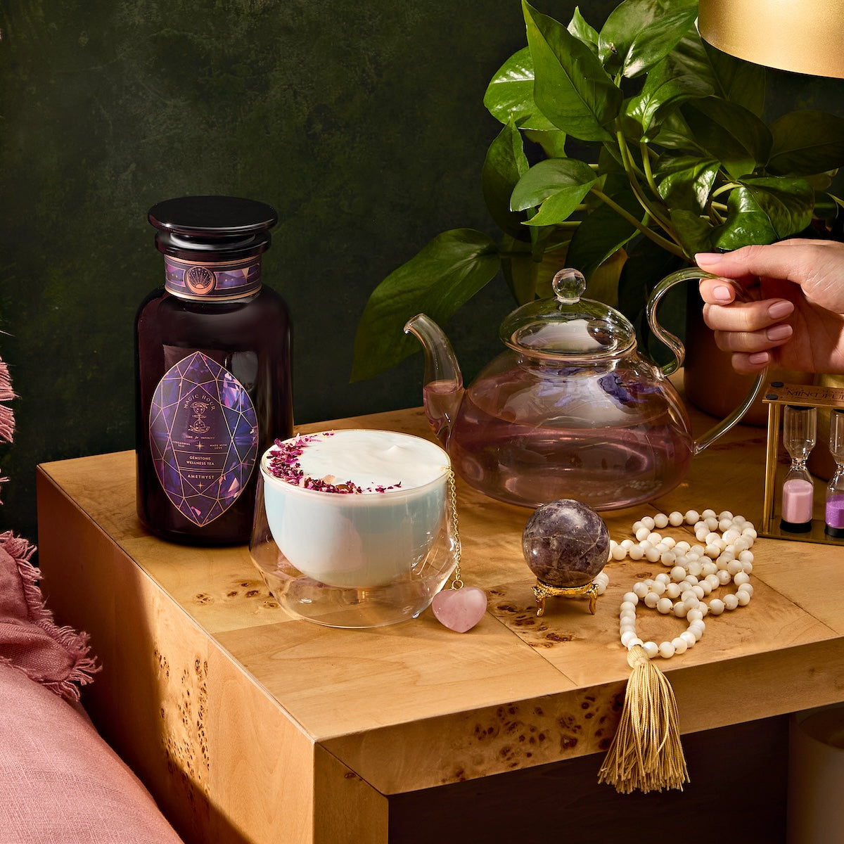 A cozy scene showcases a wooden table with Amethyst: Strawberry-Passionfruit Gemstone Wellness Tea in a glass teapot, paired with a frothy rose petal-topped drink. Complemented by an amethyst crystal and dark jar, a beaded tassel necklace accentuates the pink gem, while greenery enhances the spiritual ambiance.