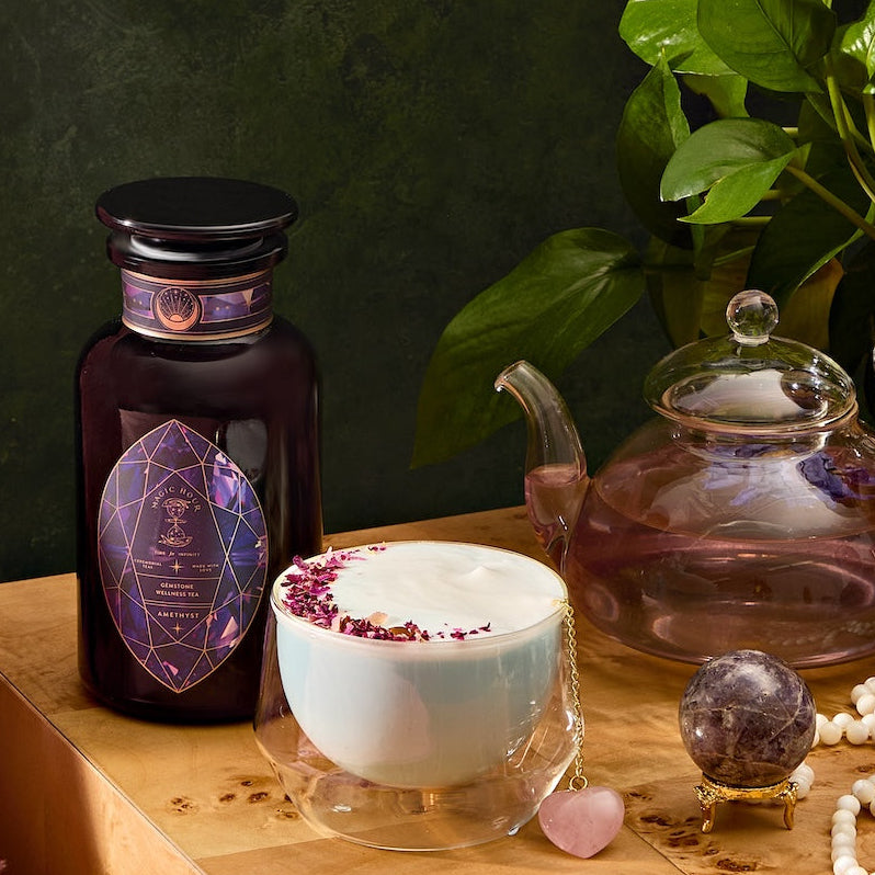 A cozy scene showcases a wooden table with Amethyst: Strawberry-Passionfruit Gemstone Wellness Tea in a glass teapot, paired with a frothy rose petal-topped drink. Complemented by an amethyst crystal and dark jar, a beaded tassel necklace accentuates the pink gem, while greenery enhances the spiritual ambiance.