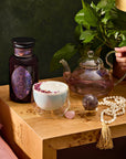 A cozy scene showcases a wooden table with Amethyst: Strawberry-Passionfruit Gemstone Wellness Tea in a glass teapot, paired with a frothy rose petal-topped drink. Complemented by an amethyst crystal and dark jar, a beaded tassel necklace accentuates the pink gem, while greenery enhances the spiritual ambiance.