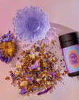 A purple translucent soap with butterfly pea flower, dried petals, and herbs sits beside a "111: Lavender-Jasmine-Pineapple Herbal Tea with Blue Lotus" container on a pink and purple gradient background.