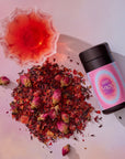 Hibiscus-Papaya Oolong with Roses loose tea leaves are scattered near a glass cup of red tea and a container labeled "222" on a soft pink background, highlighting this unique rose blend.