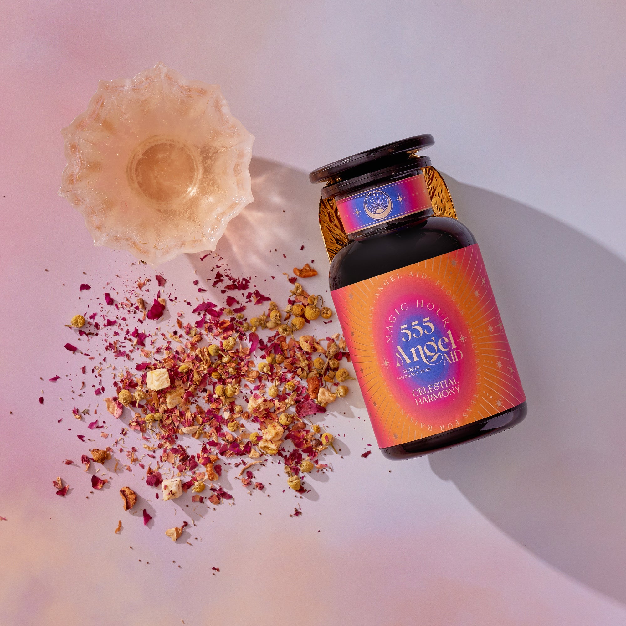 A glass jar labeled "555: Celestial Harmony: Papaya-Mango-Rose Tea with Ashwagandha" rests on a pink surface beside a dish and vibrant dried petals, evoking stress relief and the soothing magic of this unique tea blend in a dreamy, magical scene.