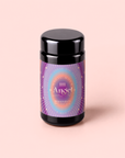 A black jar with a colorful, radiant label reading “111” and “Lavender-Jasmine-Pineapple Herbal Tea with Blue Lotus” sits on a pale pink background. The label, adorned with purple, blue, and gold decorative details, hints at the enchanting blue herbal tea within—an infusion inspired by Egyptian blue lotus and enhanced with lavender, jasmine, and pineapple notes.