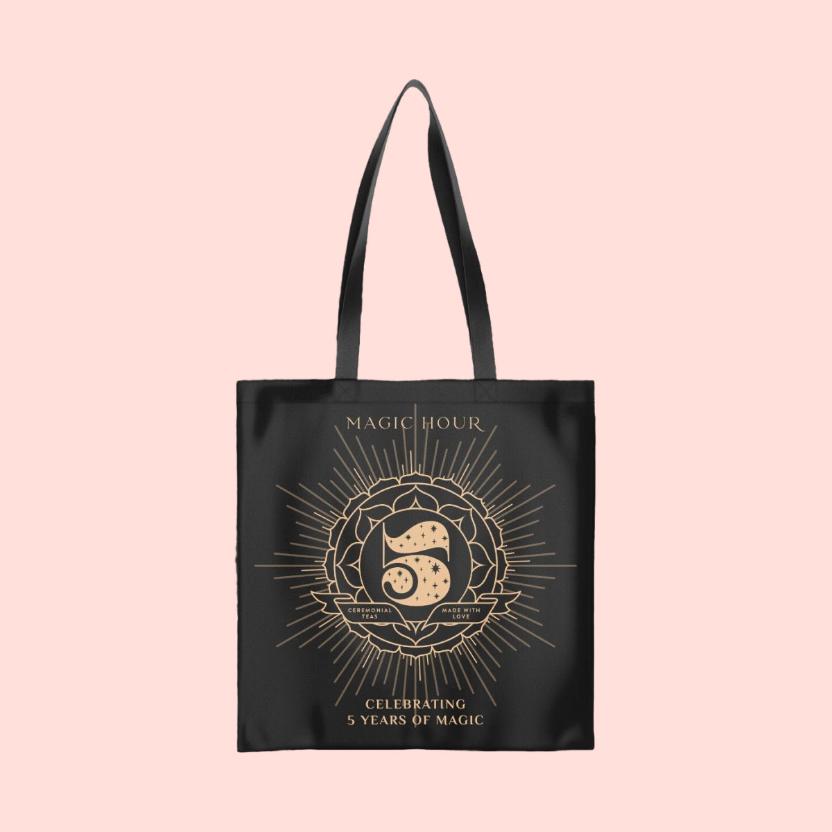 A black cotton tote bag named the Magic Hour Tote Bag showcases &quot;Magic Hour&quot; at the top, with a decorative number 5 surrounded by a floral and sunburst design. Below this, a metallic gold logo states &quot;Celebrating 5 Years of Magic,&quot; all set against a soft pink background.