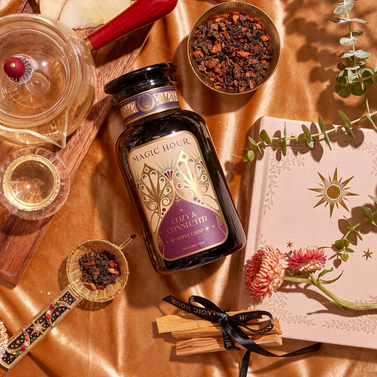 A flat lay photo features a bottle of &quot;Apple Crisp Oolong&quot; labeled &quot;Cozy &amp; Connected.&quot; Surrounding it are a teacup, a tea infuser, a wooden tray, tea leaves in bowls, a pink book with a sun design, and eucalyptus leaves on a satin golden fabric background exuding warm autumn vibes.