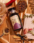 A flat lay photo features a bottle of "Apple Crisp Oolong" labeled "Cozy & Connected." Surrounding it are a teacup, a tea infuser, a wooden tray, tea leaves in bowls, a pink book with a sun design, and eucalyptus leaves on a satin golden fabric background exuding warm autumn vibes.