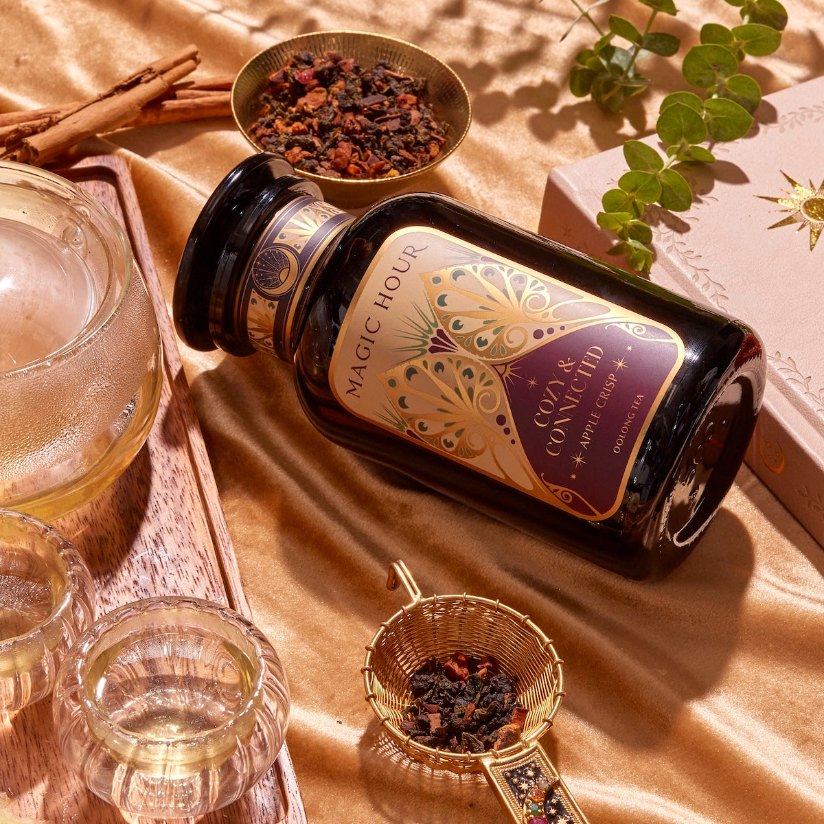 A jar labeled "Apple Crisp Oolong" lies on a gold satin fabric, evoking autumn vibes. Surrounding the jar are two glass cups, a metal strainer with loose Oolong tea leaves, a glass teapot, cinnamon sticks, and a decorative book with a green plant.