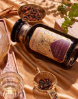 A jar labeled "Apple Crisp Oolong" lies on a gold satin fabric, evoking autumn vibes. Surrounding the jar are two glass cups, a metal strainer with loose Oolong tea leaves, a glass teapot, cinnamon sticks, and a decorative book with a green plant.