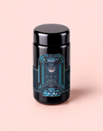 A black cylindrical container with a geometric blue label, set against a light pink background. The intricate label design and text suggest it is Aquamarine Dream – Soothing Herbal Ayurvedic Adrenal Tonic, an adaptogen sleep blend, though the finer details remain out of focus.