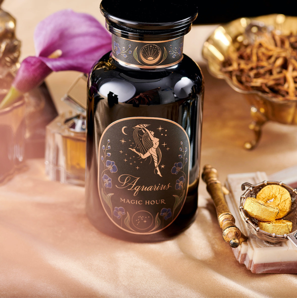 A black glass jar labeled "Aquarius: Visionary Goddess Tea" with gold and floral designs sits on gold satin, surrounded by a purple calla lily, dried herbs, gold-wrapped chocolates, and Organic Golden Monkey Tip Black Tea leaves.