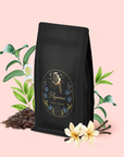 A package of "Aquarius: Visionary Goddess Tea" featuring celestial artwork is surrounded by tea leaves, white blossoms, green leaves, and vanilla flowers, hinting at an enchanting vanilla black tea blend from Yunnan, all set against a light pink background.