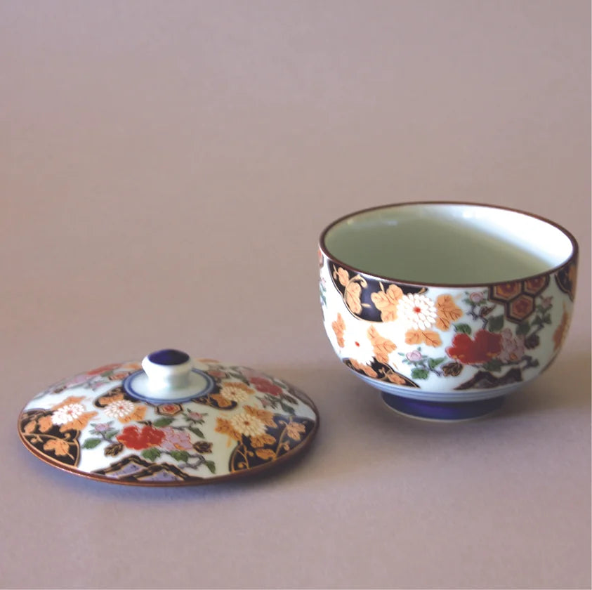 An Eastern Elm Arita Peony Flower Tea Cup with Lid, both delicately decorated with intricate floral and geometric patterns in vibrant hues of red, gold, and white. The lid is situated to the left of the tea cup. The background is a plain, neutral surface accentuating the exquisite Japanese craftsmanship.