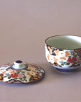 An Eastern Elm Arita Peony Flower Tea Cup with Lid, both delicately decorated with intricate floral and geometric patterns in vibrant hues of red, gold, and white. The lid is situated to the left of the tea cup. The background is a plain, neutral surface accentuating the exquisite Japanese craftsmanship.