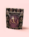 A decorative bag of "Artemis of the Wilds: Wild Moon White Tea," adorned with an illustrated archer goddess and ornate floral patterns on a pale pink background. This enchanting low-caffeine tea is perfect for savoring serene moments at any time of day.