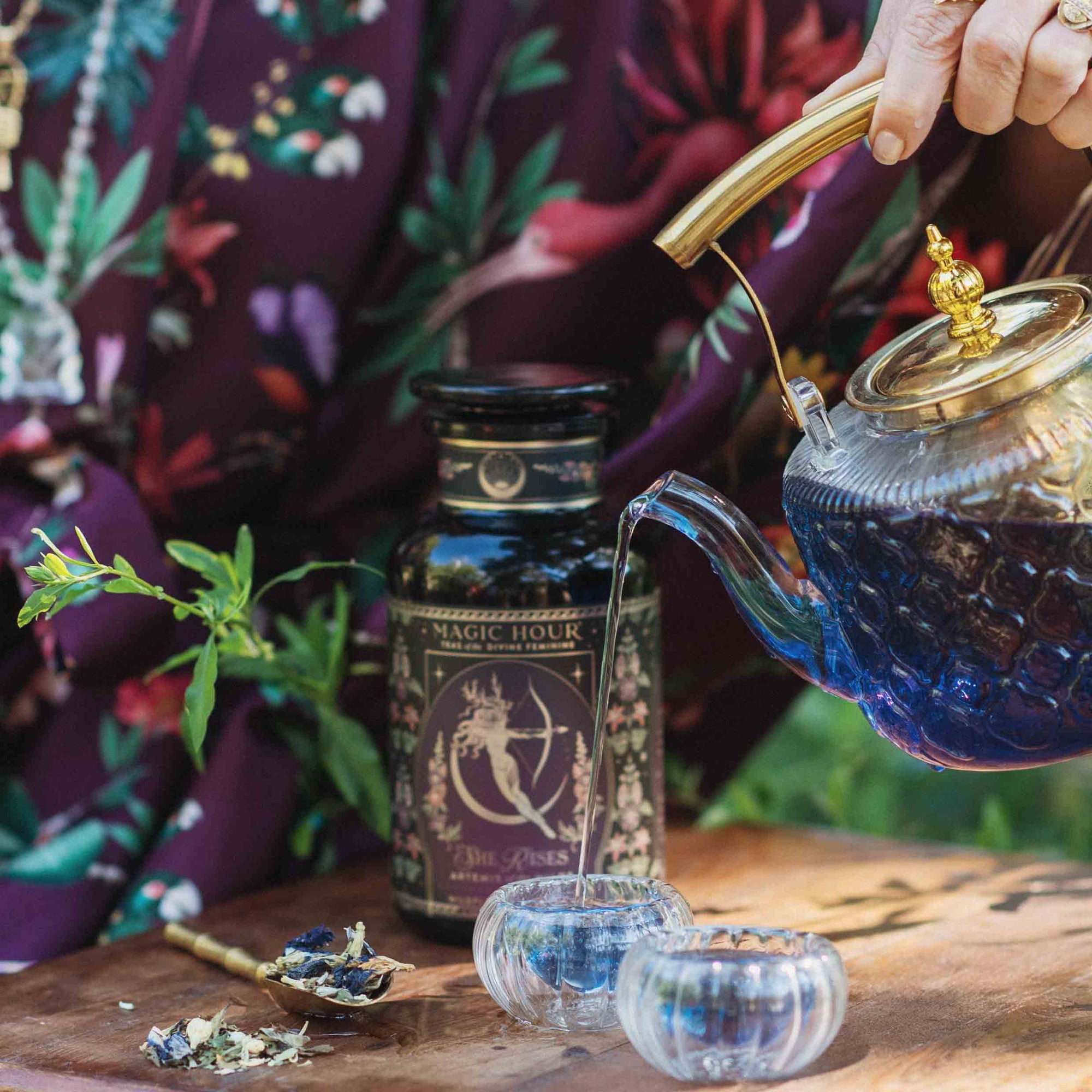 A hand pours Artemis of the Wilds: Wild Moon White Tea from a blue glass teapot into two small cups on a wooden table, with the floral tea jar and fresh herbs nearby. The person is dressed in a floral-patterned garment.