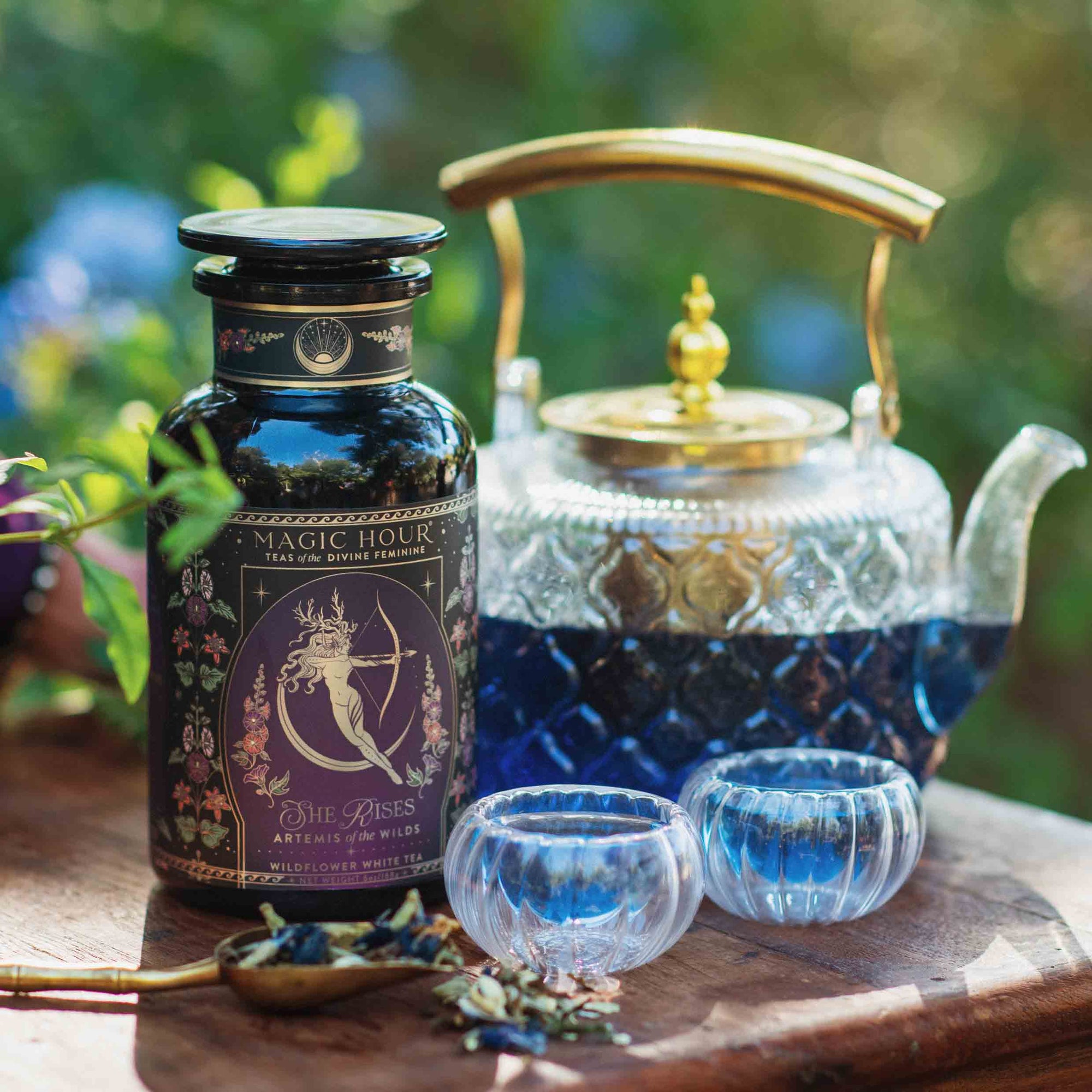 Artemis of the Wilds: Wild Moon White Tea, a captivating low-caffeine blend, is displayed in a glass teapot with two color-changing cups beside a decorative "Magic Hour" canister, set outdoors amid lush greenery.
