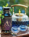 Artemis of the Wilds: Wild Moon White Tea, a captivating low-caffeine blend, is displayed in a glass teapot with two color-changing cups beside a decorative "Magic Hour" canister, set outdoors amid lush greenery.