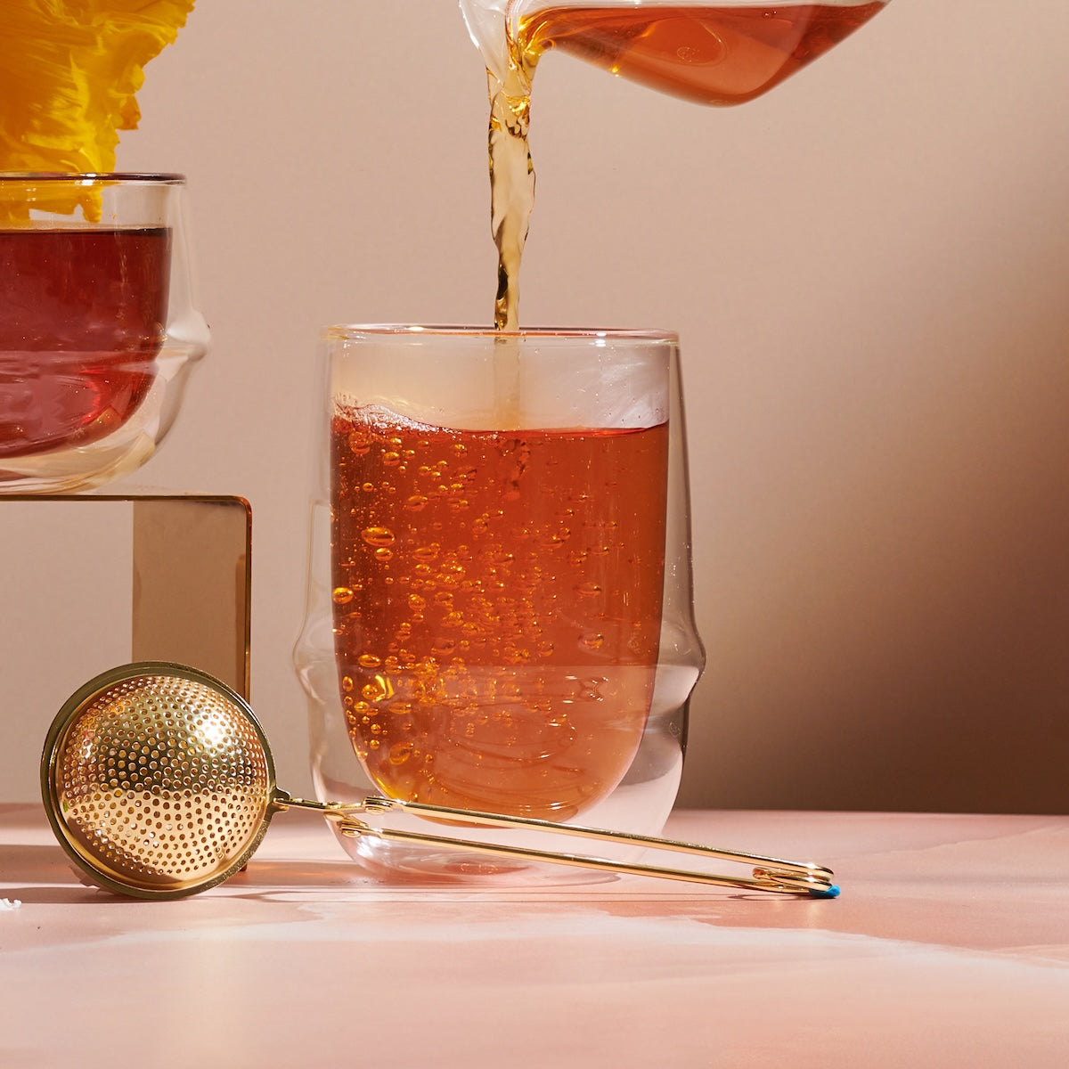 A close-up of tea being poured from a glass container into a Kinto Double-Walled Iced Tea Glass: 350mL by Magic Hour. A gold tea infuser with a long handle rests nearby on a light pink table. The setup is bright and clean, with a soft, neutral background.