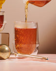 A close-up of tea being poured from a glass container into a Kinto Double-Walled Iced Tea Glass: 350mL by Magic Hour. A gold tea infuser with a long handle rests nearby on a light pink table. The setup is bright and clean, with a soft, neutral background.