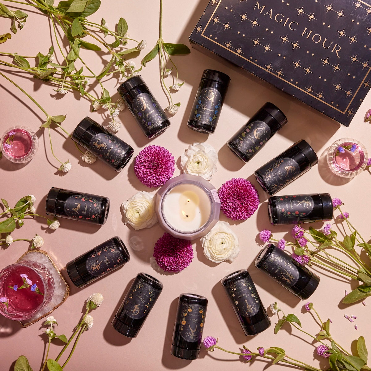 A circle of small black cylindrical bottles with intricate designs surrounds a white candle and pink and white flowers, creating a serene tea ritual. A box labeled "Astrology Tea Traveler Bundle" sits at the top, while scattered green stems add a touch of nature to the arrangement.