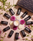 A circle of small black cylindrical bottles with intricate designs surrounds a white candle and pink and white flowers, creating a serene tea ritual. A box labeled "Astrology Tea Traveler Bundle" sits at the top, while scattered green stems add a touch of nature to the arrangement.