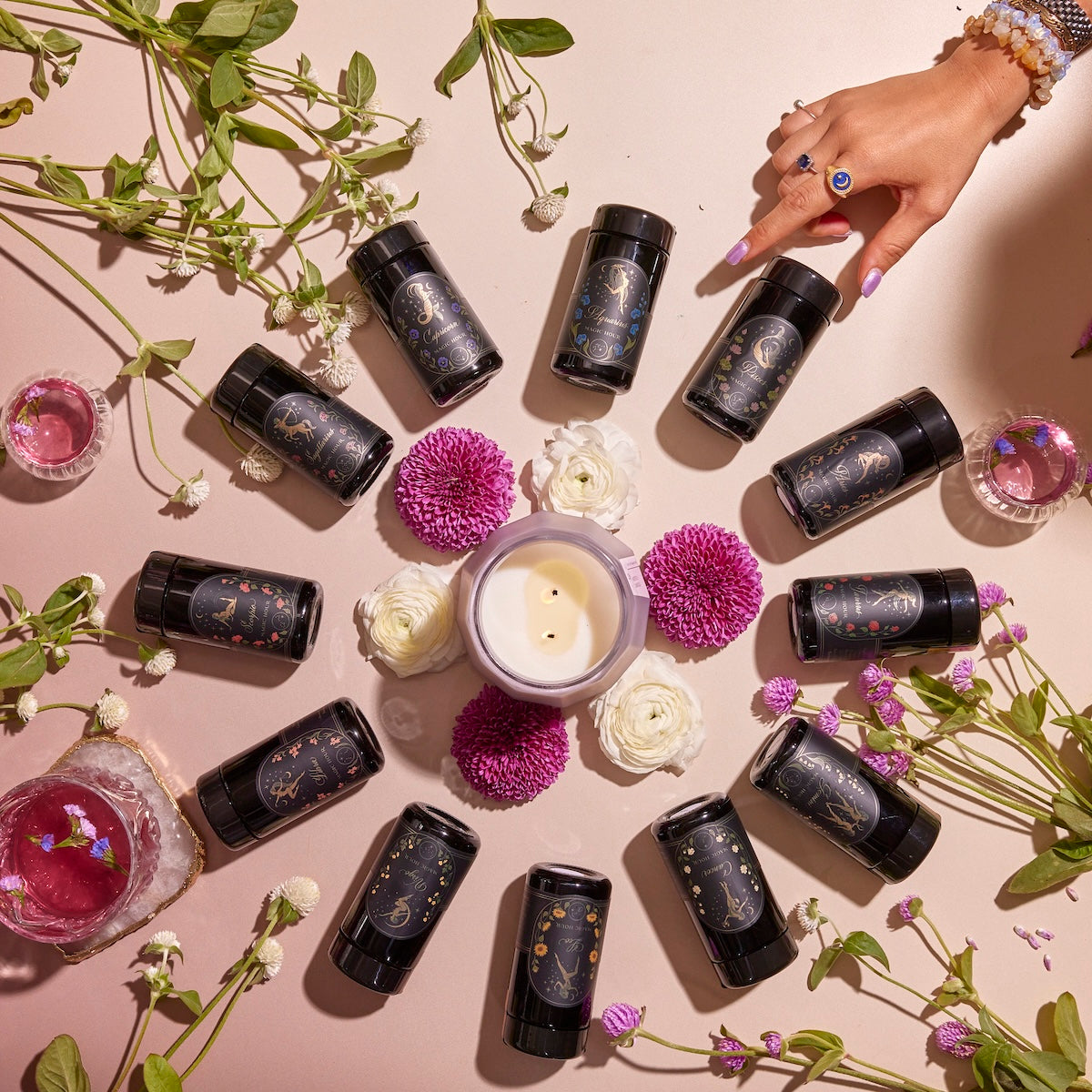 A captivating display of the Astrology Tea Traveler Bundle features black skincare bottles arranged in a circle with a lit candle and vibrant flowers at the center, evoking the calming essence of a tea ritual. A hand adorned with purple nail polish and bracelets highlights one bottle, while scattered greenery enhances the fresh vibe on the soft pink background.