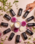 A captivating display of the Astrology Tea Traveler Bundle features black skincare bottles arranged in a circle with a lit candle and vibrant flowers at the center, evoking the calming essence of a tea ritual. A hand adorned with purple nail polish and bracelets highlights one bottle, while scattered greenery enhances the fresh vibe on the soft pink background.